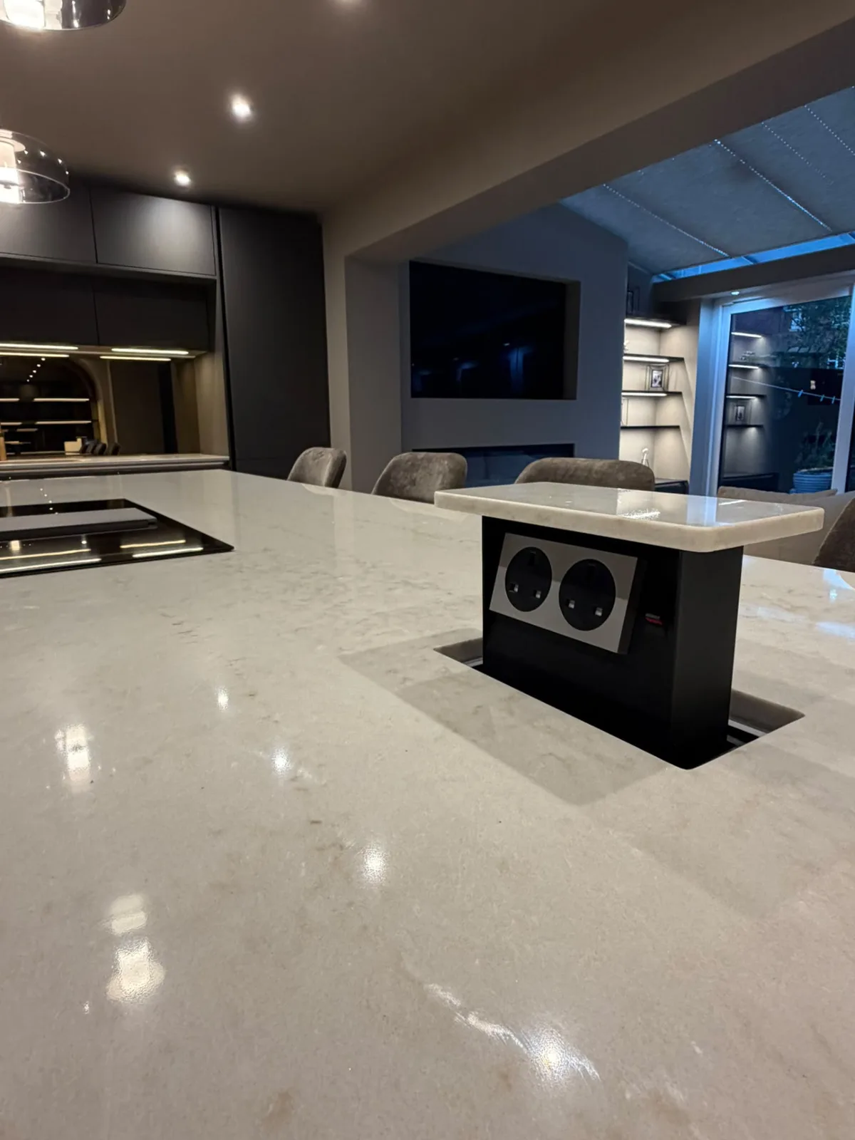 Pop-up power sockets built into a kitchen island worktop with recessed ceiling downlights and under-cabinet LED strip lighting